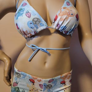 May include: A light blue bikini set with a vibrant print of cartoon characters and berries. The set comprises a triangle top with thin straps and a matching low-rise bottom. The bikini is displayed on a mannequin.