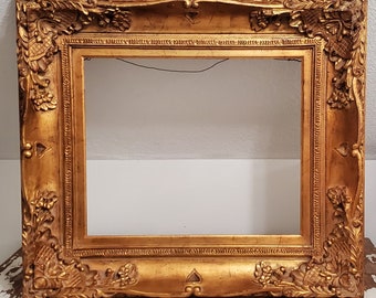 May include: Ornate gold picture frame with detailed floral and scrollwork carvings. The frame has a rectangular opening and a rich, aged appearance. The frame is made of wood and is suitable for artwork or photographs.
