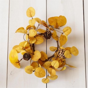 May include: A circular wreath featuring yellow leaves and small brown pinecones. The leaves are arranged in a dense pattern, creating a full and textured appearance. The wreath is set against a white background.
