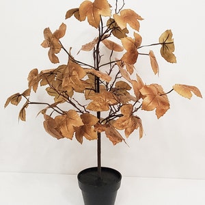 May include: A small artificial tree with brown leaves in a black pot. The tree is made of plastic and has a realistic look.