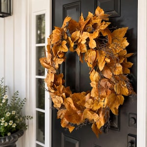 May include: A circular wreath composed of numerous golden-brown autumn leaves and thin, dark brown twigs. The foliage features varied shades of amber and tan, creating a dense, layered texture throughout the round arrangement of dried leaves.