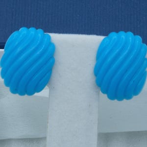 May include: A pair of bright blue, oval-shaped earrings with a swirled design. The earrings are displayed in a white box with a blue lid. The earrings are a vibrant shade of blue.