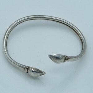 May include: A silver-colored open cuff bracelet. The bracelet has a smooth, rounded band that tapers to pointed ends. The ends have a decorative detail near the tip. The bracelet is designed to be worn on the wrist.