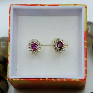 May include: A pair of gold stud earrings, each featuring a central oval-cut red gemstone surrounded by a halo of small clear stones. The earrings are presented inside a square box with a red and yellow patterned exterior and white interior.