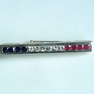 May include: A silver tie bar featuring a line of gemstones in blue, clear, and red. The gemstones are set in a rectangular bar. The design is simple and elegant, suitable for formal wear.