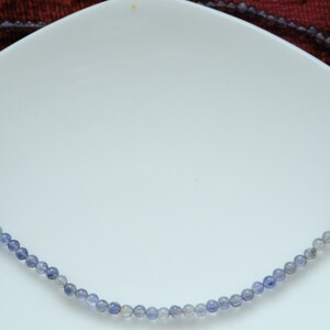 May include: A delicate necklace made of small, round, blue and gray beads. The necklace is on a white surface.