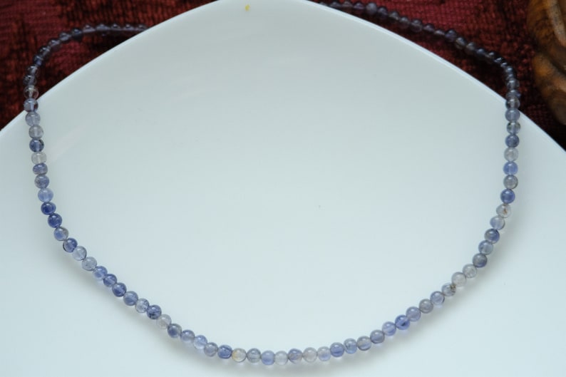May include: A delicate necklace made of small, round, blue and gray beads. The necklace is on a white surface.
