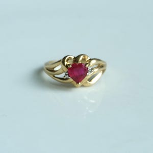 Puede incluir: Un anillo de oro con una piedra preciosa roja oscura en forma de corazón. El anillo tiene un diseño de caña dividida con pequeños diamantes de acento. Una pieza de joyería clásica.