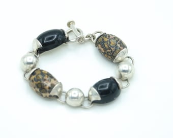 May include: A silver bracelet featuring a toggle clasp. The bracelet has a series of beads, including black, speckled brown, and silver spheres. The beads are connected by silver links.
