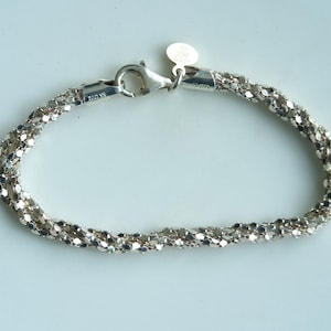 May include: A silver-colored bracelet with a twisted rope design. The bracelet has a lobster clasp closure and a small, round tag. The jewelry is made of a shiny, metallic material and is displayed on a light-colored surface.