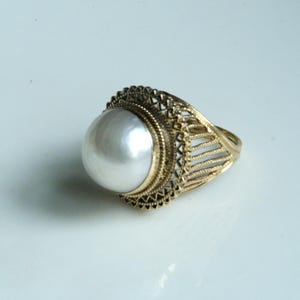May include: A gold-toned ring featuring a large, round, white pearl. The ring has an ornate, filigree design around the pearl and on the band. The ring is set against a white background.