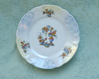 May include: A white, scalloped-edge plate with floral designs. The plate features a central bouquet of flowers in a vase, with additional floral clusters around the rim. The design includes orange, blue, and yellow hues.