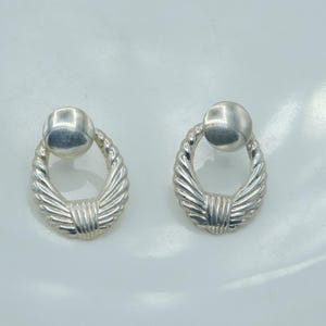 Könnte beinhalten: Ein Paar silberfarbene Ohrstecker. Jeder Ohrring hat einen kleinen, runden Stecker, der mit einem strukturierten, ovalen Design verbunden ist. Das Oval hat eine gedrehte, seilartige Textur an den Rändern.