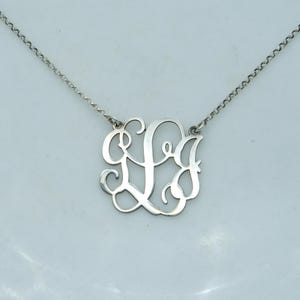 以下が含まれることがあります： 繊細なチェーンが付いたシルバーカラーのモノグラムネックレス。ペンダントは筆記体スタイルで絡み合った文字が特徴です。ネックレスは明るい背景に表示され、ジュエリーの複雑なデザインを際立たせています。