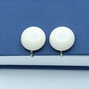 May include: A pair of white, round clip-on earrings. The earrings are set against a textured, dark blue surface. The earrings have a glossy finish and a simple, classic design, suitable for everyday wear.