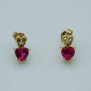 May include: A pair of gold stud earrings featuring a heart-shaped, vibrant pink gemstone suspended from a small gold heart-shaped setting. Each earring includes a tiny, clear accent stone embedded at the top of the gold heart structure.