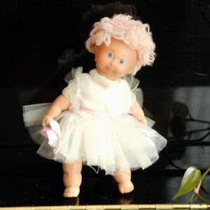 以下が含まれることがあります： ピンクのウィッグと青い目をしたヴィンテージドール。白いチュールのドレスにピンクのリボンが付いています。人形はピンクの花を持っています。人形は黒い表面に立っています。