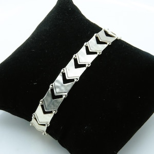 May include: A silver-toned metal bracelet featuring a series of connected chevron-shaped links. Each link has a smooth, reflective surface with a distinct V-shaped design that repeats along the length of the chain, creating a structured pattern.