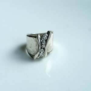 May include: A silver ring with a textured band and a row of clear stones set in the center.