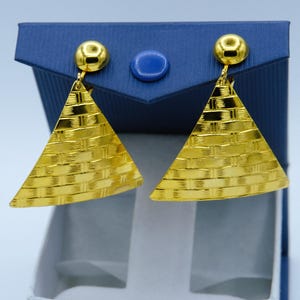 May include: A pair of gold-colored earrings with a triangular shape and a woven texture. Each earring has a small, round stud at the top. The earrings are displayed in a blue jewelry box with a white interior.
