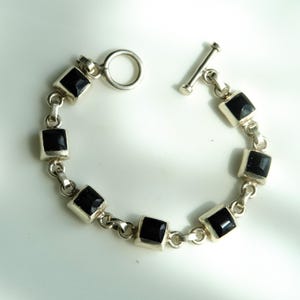 May include: Silver bracelet featuring square black gemstones. The bracelet has a toggle clasp and is composed of interconnected links. The gemstones are set in a silver metal setting. The bracelet is displayed on a white surface.