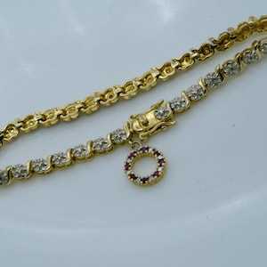 May include: A gold-coloured bracelet featuring a repeating pattern of diamond-like stones. A small, circular charm, set with red stones, dangles from the bracelet. The bracelet is on a white surface.