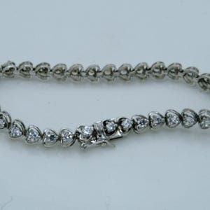 Puede incluir: Una pulsera plateada con eslabones en forma de corazón, cada uno engastado con una pequeña gema transparente. La pulsera tiene un cierre de broche seguro. La joya se muestra sobre un fondo claro.