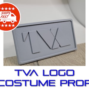 May include: A gray plastic TVA logo prop for cosplay or costume use. The logo is a stylized "TVA" with a raised, textured design.