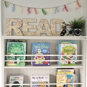 May include: A white bookshelf with wooden letters spelling "READ" above it. The bookshelf is filled with colorful children's books with titles like "General Knowledge", "See Inside Your Body", "Colors", "My 'q' Book", and "If You Take A Mouse To School".
