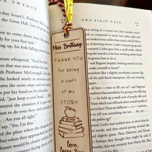 May include: A wooden bookmark with a brown background and a yellow ribbon. The bookmark features a stack of books with an apple on top, and the text "Thank you for being a part of my story!"  The bookmark also includes the text "Love, Lucas Bess".