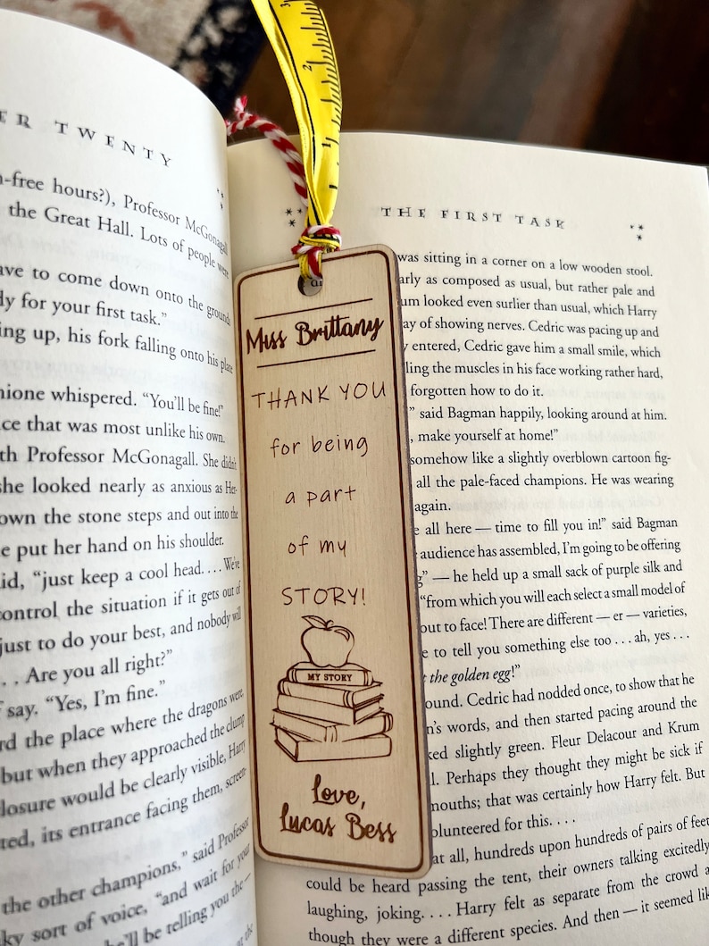 May include: A wooden bookmark with a brown background and a yellow ribbon. The bookmark features a stack of books with an apple on top, and the text "Thank you for being a part of my story!"  The bookmark also includes the text "Love, Lucas Bess".