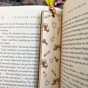 May include: A wooden bookmark with a laser-engraved design of school supplies, including a bus, scissors, pencils, glue, and hearts. The bookmark is a light brown color and has a red and white ribbon attached to it.