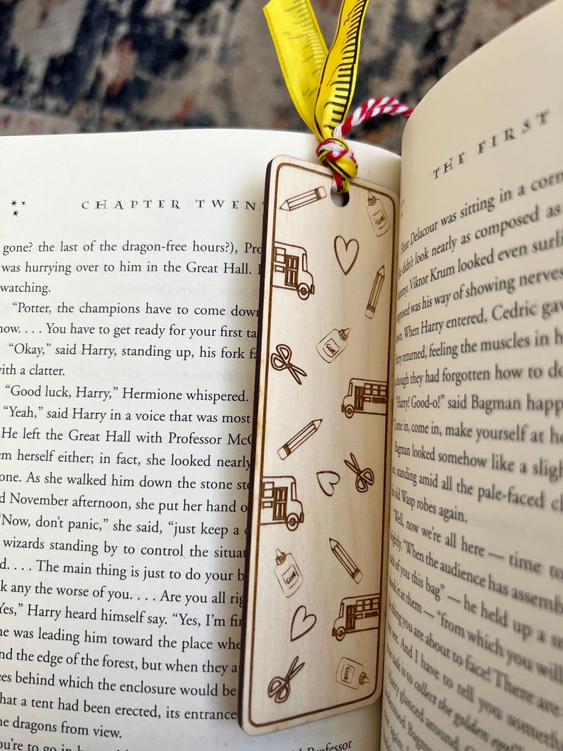 May include: A wooden bookmark with a laser-engraved design of school supplies, including a bus, scissors, pencils, glue, and hearts. The bookmark is a light brown color and has a red and white ribbon attached to it.