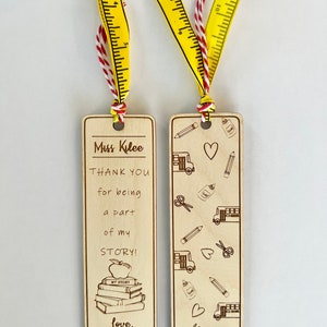May include: Two wooden bookmarks with a teacher appreciation theme. One bookmark has a stack of books with an apple on top and the text "Miss Kilee THANK YOU for being a part of my STORY! love, Lucas Bess". The other bookmark has a pattern of school busses, pencils, scissors, hearts, and glue bottles.