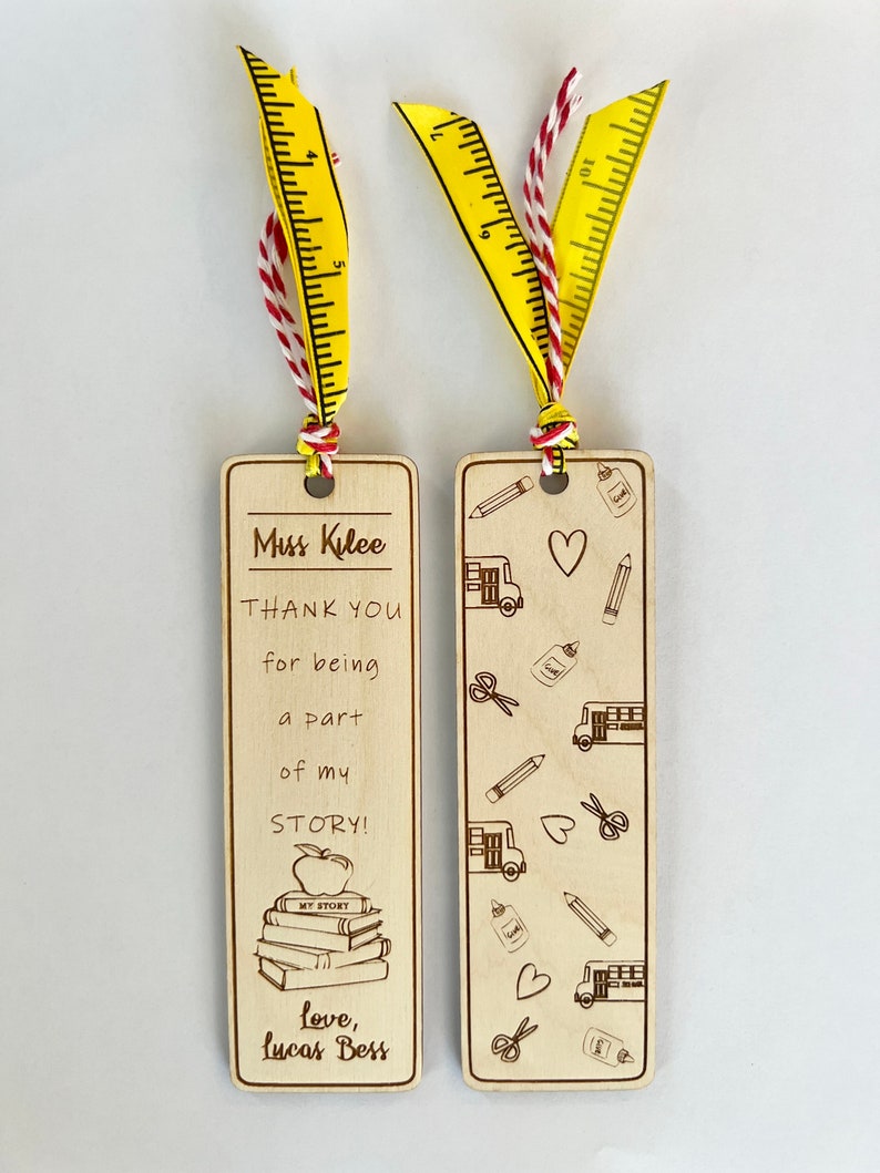 May include: Two wooden bookmarks with a teacher appreciation theme. One bookmark has a stack of books with an apple on top and the text "Miss Kilee THANK YOU for being a part of my STORY! love, Lucas Bess". The other bookmark has a pattern of school busses, pencils, scissors, hearts, and glue bottles.
