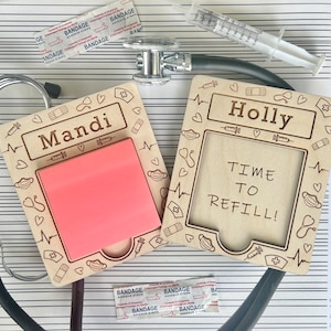 May include: Two wooden note holders with personalized names "Mandi" and "Holly". Each holder has a space for a sticky note and features a medical theme with hearts, stethoscopes, and bandages. The "Holly" holder has the text "Time to Refill!" on it.