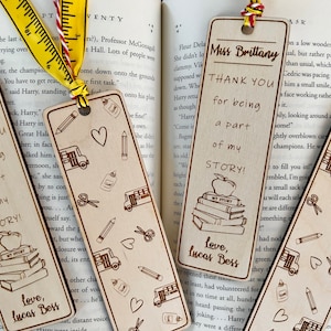 May include: Three wooden bookmarks with a teacher appreciation theme. Each bookmark features a different design with a stack of books, an apple, and the text "Thank You for being a part of my story!"  The bookmarks are decorated with hearts, pencils, scissors, and school buses.