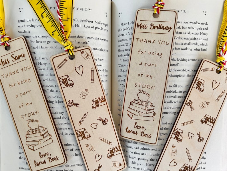 May include: Three wooden bookmarks with a teacher appreciation theme. Each bookmark features a different design with a stack of books, an apple, and the text "Thank You for being a part of my story!"  The bookmarks are decorated with hearts, pencils, scissors, and school buses.