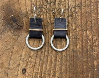 May include: A pair of handmade earrings featuring black rectangular leather pieces with a thin gold-colored band and a silver-colored hoop. The earrings hang from silver-colored ear wires. The earrings are displayed on a wooden surface.