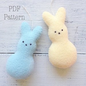 Peut inclure: Deux peluches de lapin en feutre, l'une bleue et l'autre jaune, avec des yeux noirs et un simple sourire cousu. Les lapins sont suspendus à une surface en bois blanc.