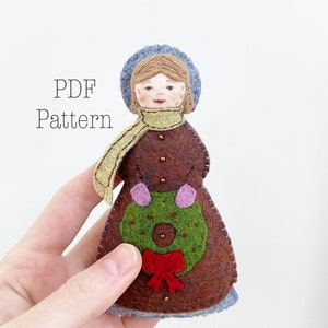May include: A felt Christmas ornament of a woman wearing a brown dress, a blue hat, and a yellow scarf. She is holding a green wreath with a red bow.
