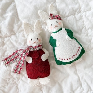 Two felt rabbit Christmas ornaments rest on a white background. The boy rabbit wears red overalls with a plaid scarf. The girl rabbit wears a green dress, white apron, and plaid bow. They are festive felt Christmas ornaments.
