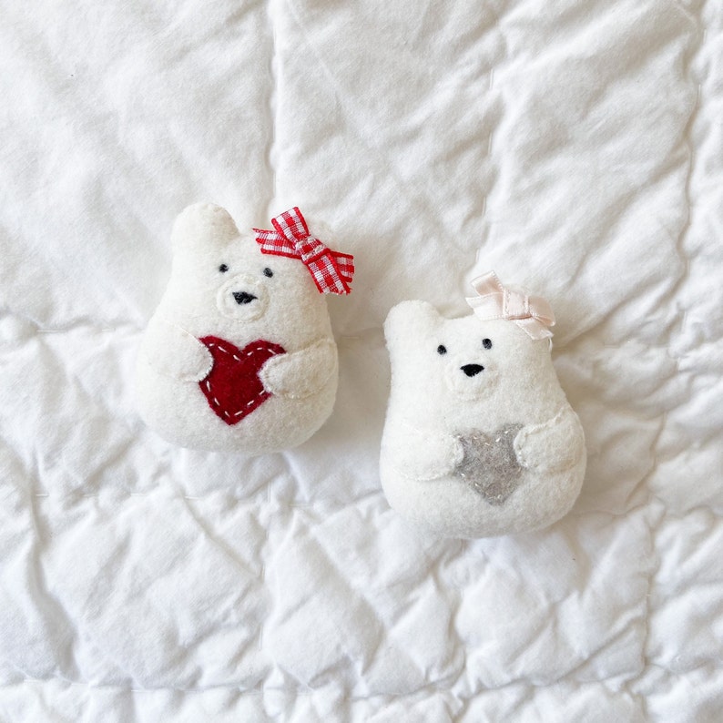 Two felt bear ornaments rest on a white fabric background. Each bear ornament is holding a heart. The bears have bows on their ears. Christmas ornament pattern. Valentine ornaments.