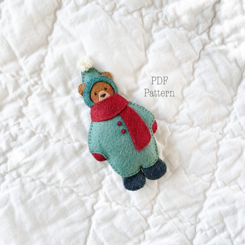 Felt Christmas Ornament. Felt Bear Ornament. Felt Christmas Bear Ornament on a white background. The bear is wearing a green felt snowsuit with a red felt scarf. The text says PDF Pattern. It is a Christmas sewing pattern.