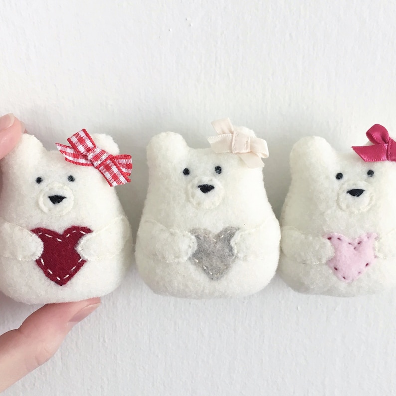 Three tiny felt bear ornaments on a white background. Each bear is holding a heart and have bows on their ears. Christmas ornament pattern. Valentine ornament pattern. Felt ornament pattern.