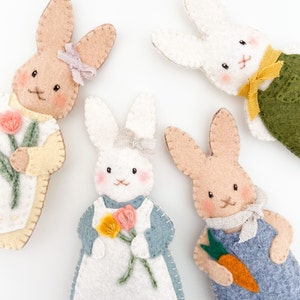 Four felt Christmas ornaments are in a circle. They are felt rabbit ornaments. Two of the rabbits are wearing dresses and holding felt flowers, and the other two rabbits are wearing overalls and holding carrots.