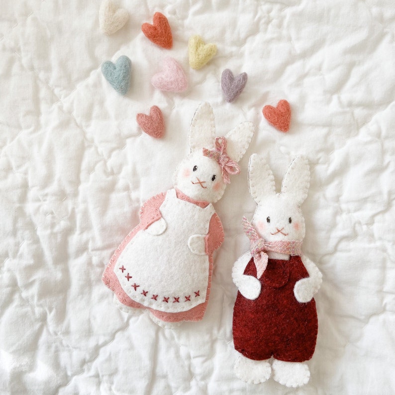 Two white rabbit felt ornaments are surrounded by colorful pastel hearts. The bunnies are dressed in valentine colors. The left bunny is white and wears a pink felt dress. The right bunny is white and wears red felt overalls. Felt Ornament Pattern.