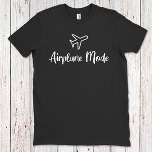 Op de afbeelding: Zwart T-shirt met een wit vliegtuigpictogram en de tekst "Vliegtuigmodus" in wit.