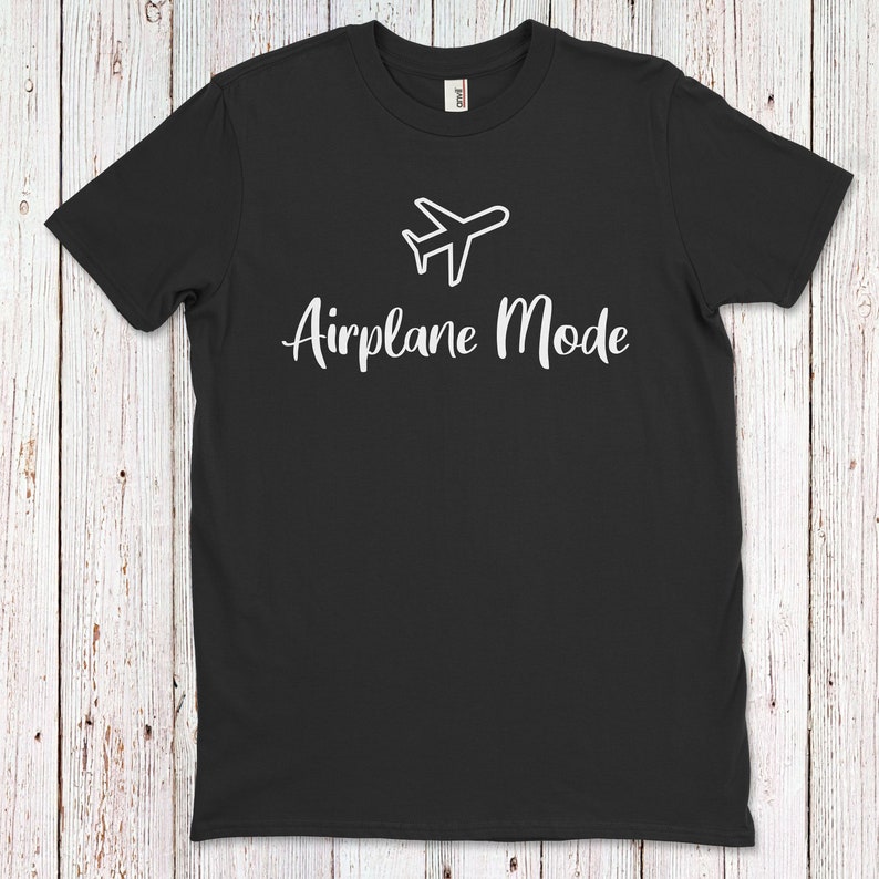 Op de afbeelding: Zwart T-shirt met een wit vliegtuigpictogram en de tekst "Vliegtuigmodus" in wit.