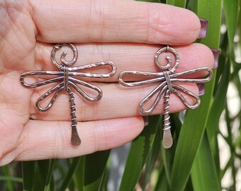 Puede incluir: Un par de pendientes de libélula de color cobre. Cada pendiente presenta un diseño de libélula con alas curvas, una cabeza en espiral y un cuerpo texturizado. Los pendientes se sostienen sobre un fondo de follaje verde.